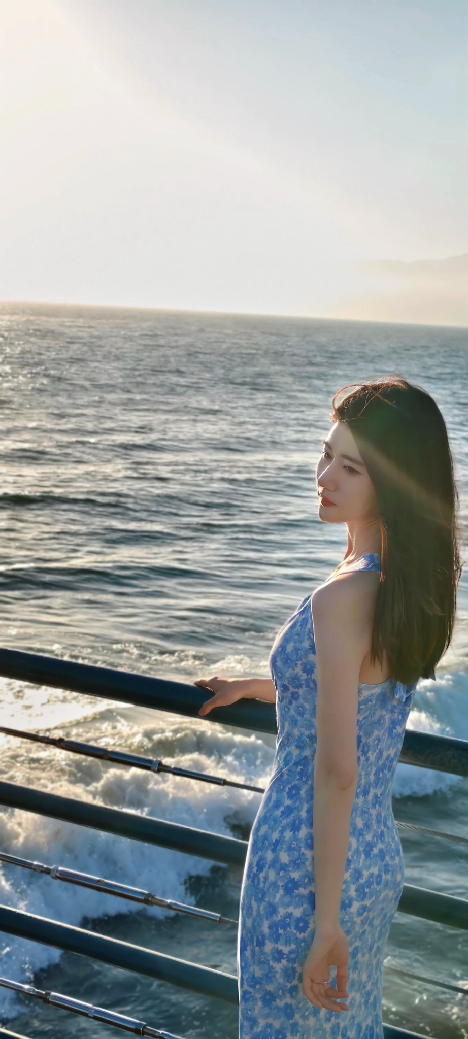 Celina in a blue floral dress beside the ocean in warm sunlight